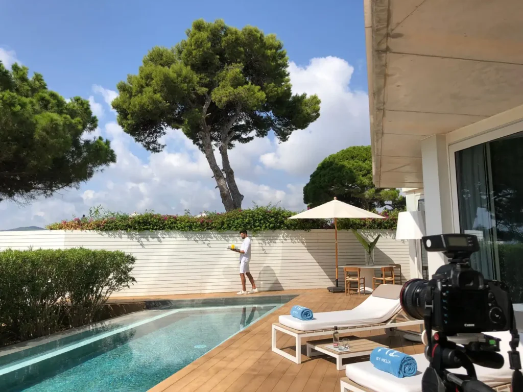 Behind the scenes of a hotel shoot: camera on a tripod facing a poolside terrace and a pine tree