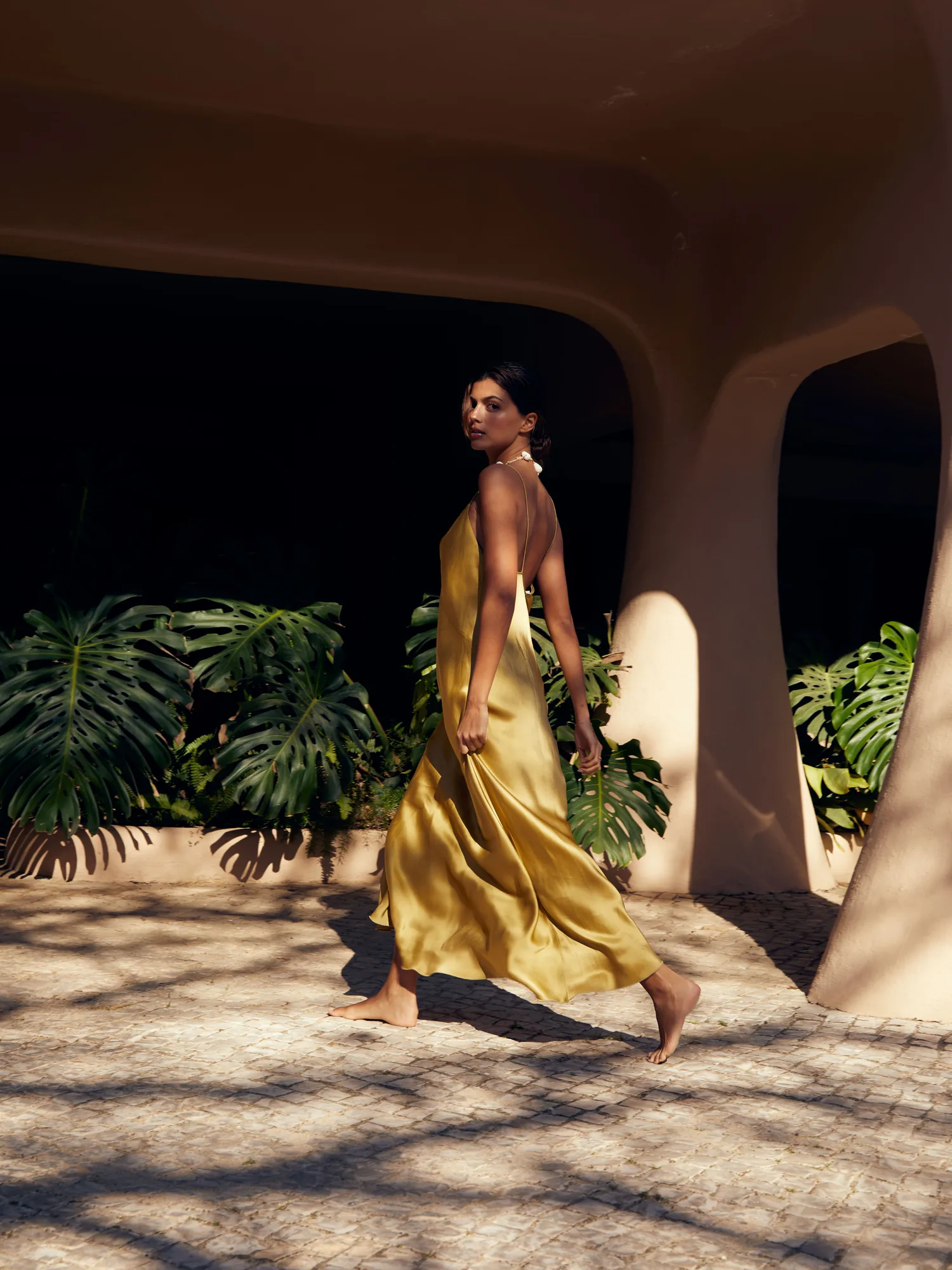 PSC-EuSouLara-golden-slip-dress · Permanent State of Creation A woman in a golden silk slip dress walking barefoot beneath the sculpted arches of Vilalara