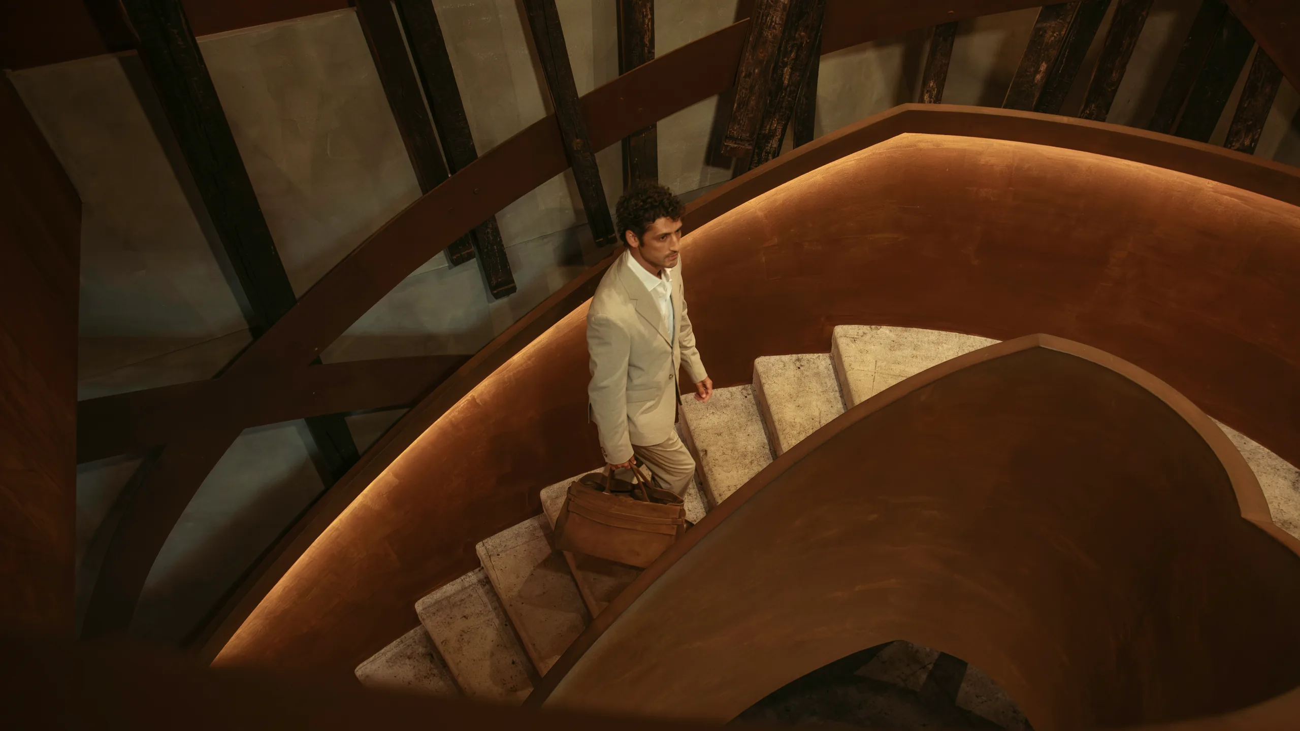 A man in a light suit carrying a leather bag climbs a spiral staircase, from The First Emotion campaign