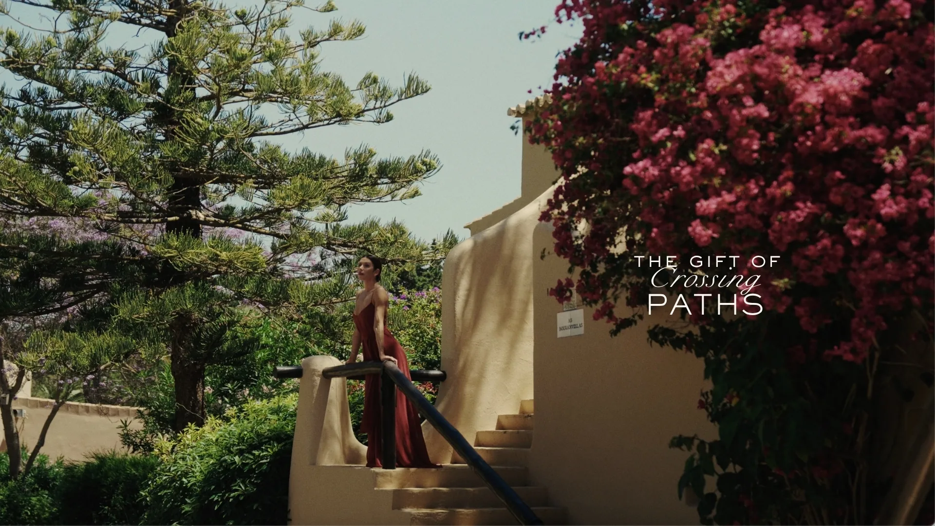 The Gift of Crossing Paths: a woman in a red dress on the whitewashed steps of Vilalara, framed by bougainvillea and pines